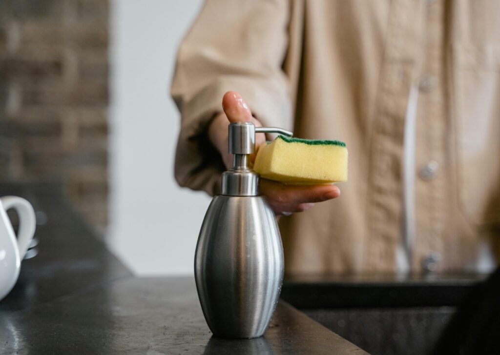 Close-up of a person using a metal soap dispenser with a cleaning sponge indoors.