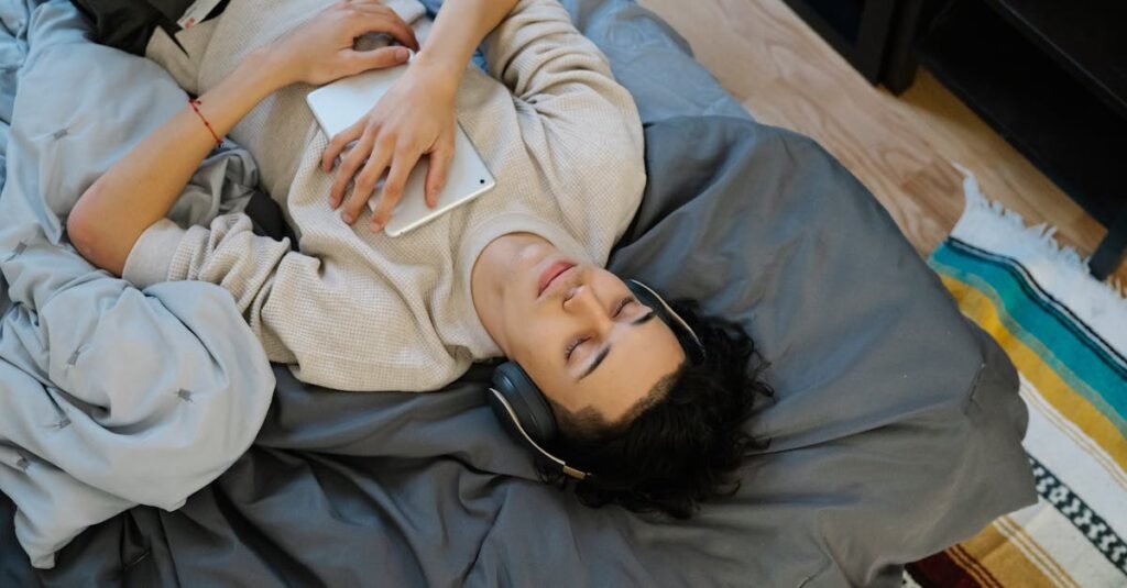 From above young Hispanic guy with tablet listening to music with closed eyes while chilling on comfortable bed at home